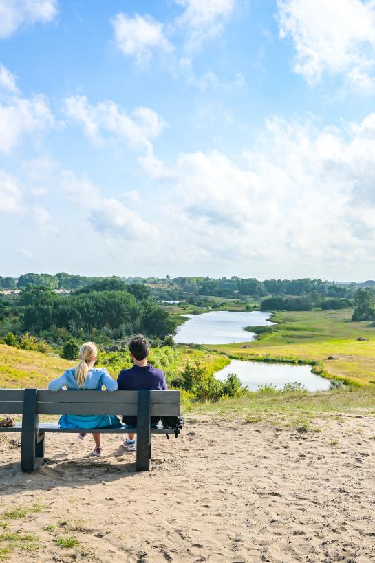 Een koppel rust uit op een bankje met zicht op een duinenlandschap